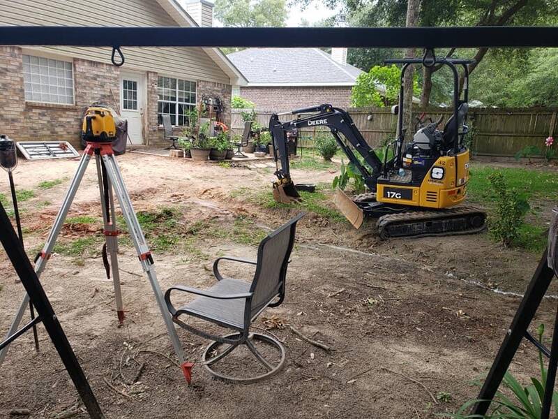 Laser level on a tripod in a bare-dirt backyard, with a yellow mini excavator by the patio of a brick house