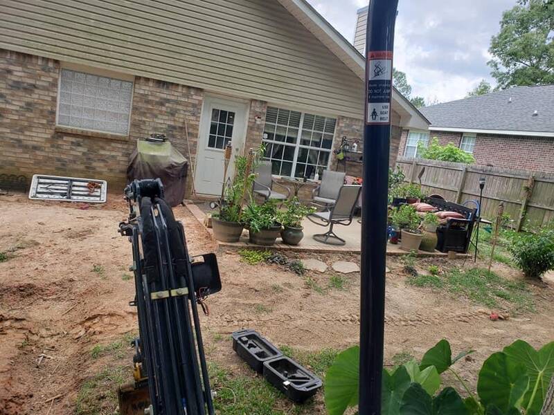 Bare-dirt backyard by a brick house patio, with drain parts and a grate laid out and an excavator arm in the foreground