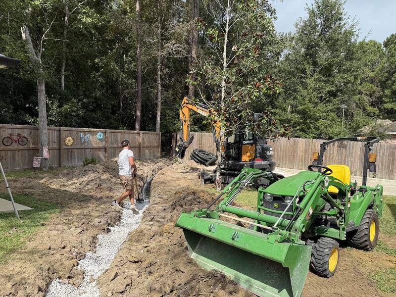 Backyard drain trench lined with white gravel, with black corrugated pipe and a mini excavator beside it